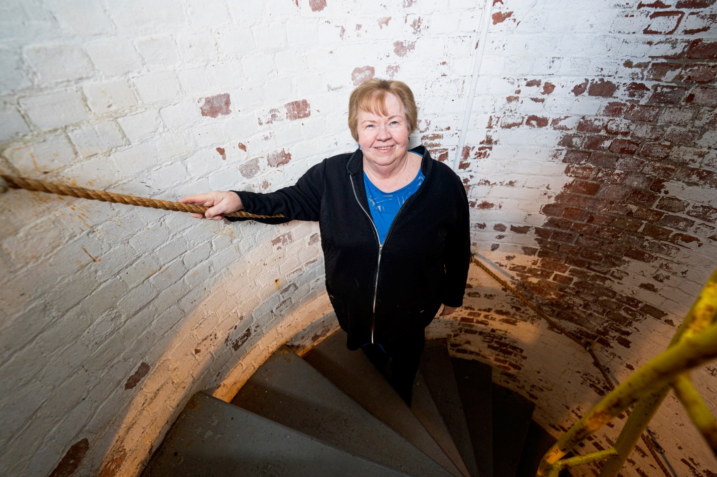 A color photograph of a person standing on a spiral staircase inside a brick-walled structure, holding a rope handrail. No readable text is visible.