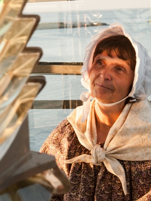 A portrait-style photo of a woman wearing a bonnet and shawl, lit by warm sunlight, with a lighthouse lens or glass structure in the foreground and water visible in the background. No readable text is present.