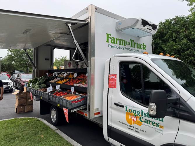 A photo of a Long Island Cares “Farm to Truck” mobile food truck with its side open, showing crates of fresh produce. The truck has prominent printed text/logos (e.g., “Farm to Truck,” “Long Island Cares,” and related branding) that could be transcribed.