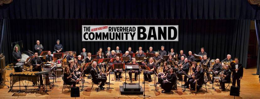 A wide group photo of a concert band seated on a stage with instruments. There is prominent banner text across the top reading “THE RIVERHEAD COMMUNITY BAND,” which is worth transcribing.