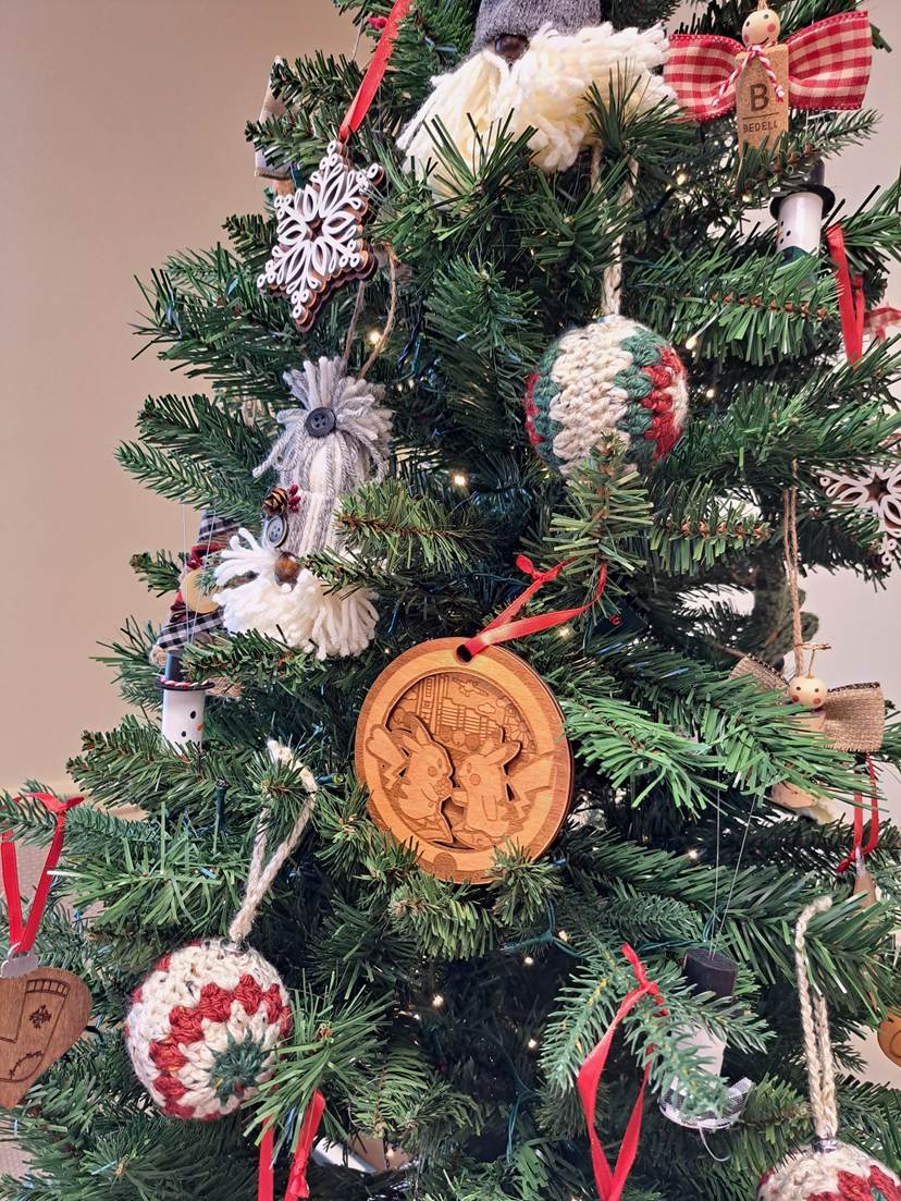 A close-up photo of a decorated Christmas tree with various handmade ornaments (crocheted balls, wooden ornaments, ribbons). There is a small tag with a letter “B” visible, but no substantial text that needs transcription.